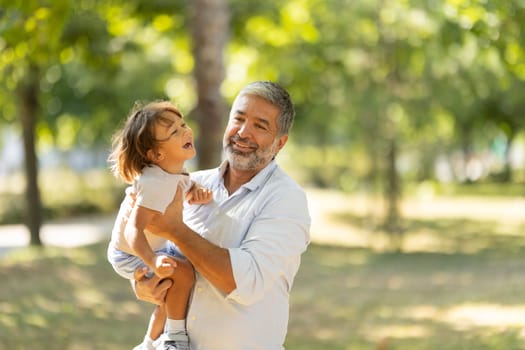 Grandfather is holding his grandson in a park and they are both smiling on a sunny day