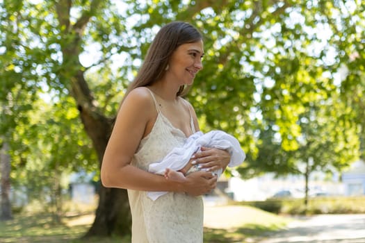 Young mother is smiling while cradling her newborn baby in a lush green park