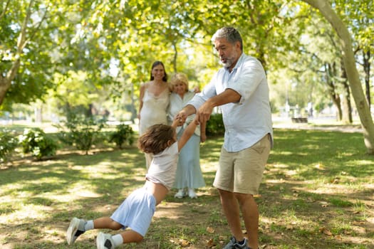Grandfather is swinging his young grandson in the air while the rest of the family watches in amusement