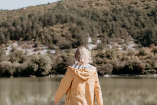 A woman wearing a yellow raincoat stands by a lake, facing away from the camera, with a lush green hillside in the background.