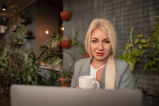 A blonde woman is sitting at a table with a laptop and a white coffee cup. She is wearing a gray jacket and has red lipstick on. The scene suggests a casual and relaxed atmosphere