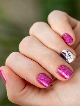 Female hand with pink manicure. Woman fingernails with pink holographic nail polish lacquer on green lush background. Aesthetic visual of red violet manicure with lady silhouette stamping nail art