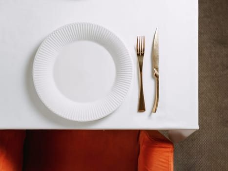 Top view of table in restaurant or dinning room with white round empty plate and gold cutlery. White tablecloth, red chair and table setting as background for your text or design. Copy space