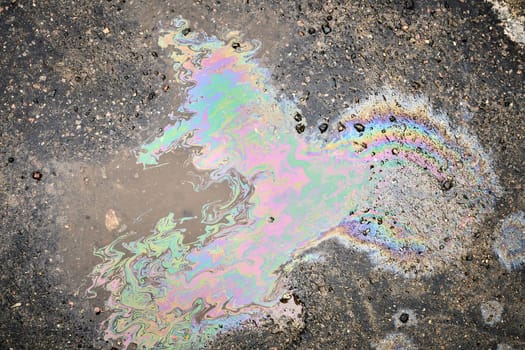 A close-up of a shimmering oil or fuel spill on wet asphalt, viewed from above.
