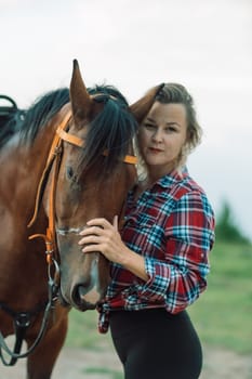 Happy blonde with horse in forest. Woman and a horse walking through the field during the day. Dressed in a plaid shirt and black leggings