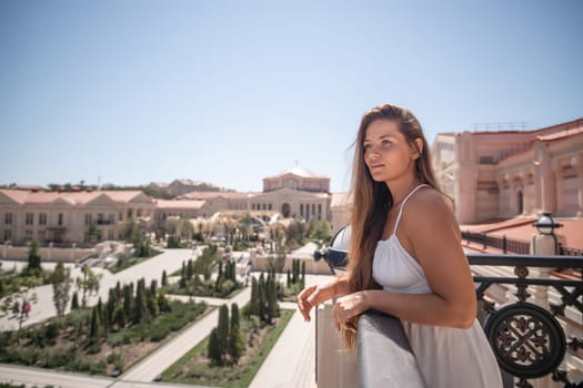 A woman in a white dress stands on a balcony overlooking a sprawling cityscape. Buildings with terracotta roofs and manicured gardens are visible in the distance. The sky is clear and blue.