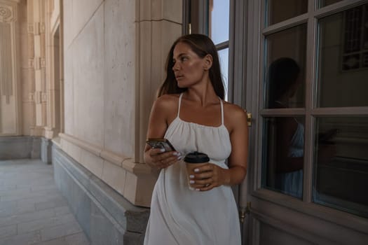 A woman in a white dress stands in a doorway, holding a coffee cup and a phone. She looks to her left. The doorway is framed by a large window.