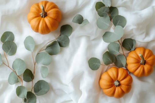 Three vibrant orange pumpkins are artfully arranged alongside eucalyptus leaves on a soft, white fabric, creating a cozy autumn atmosphere perfect for seasonal decor.
