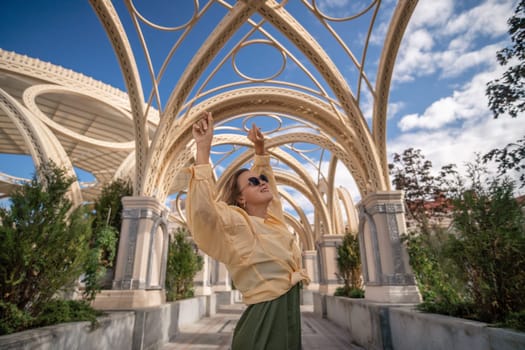 A woman in a yellow sweater and green skirt dances under a large, intricate golden structure. The sky is blue and there are clouds in the background.
