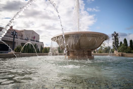 A large stone fountain with water spraying upwards in multiple streams. The fountain is surrounded by green foliage and a building in the background.