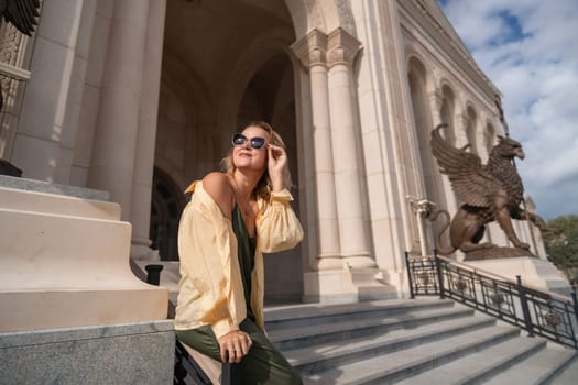 A woman in sunglasses sits on the steps of a grand building, adjusting her glasses with a smile. Two large sculptures are visible on the right.