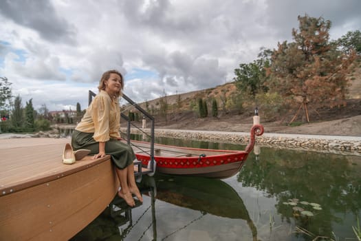 A woman sits on a wooden dock overlooking a still lake. A red boat is tied to the dock.