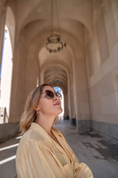 A woman wearing sunglasses and a yellow shirt stands under an archway looking up. The archway is made of stone and has a decorative chandelier hanging from the ceiling.
