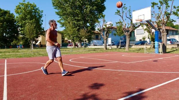 An elderly old Italian man plays basketball on a sports court on a summer day.