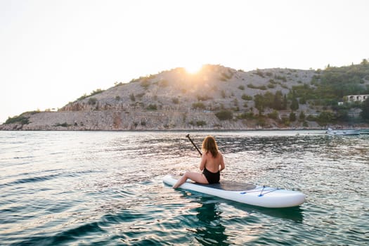 Woman paddle boarding at sunset on tranquil water near a mountain shoreline.