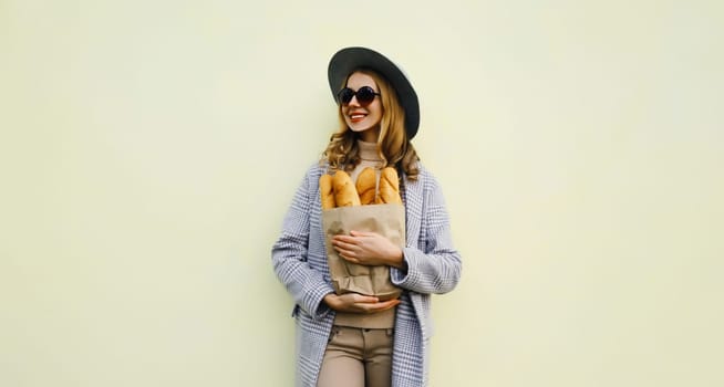 Portrait of beautiful happy smiling young woman holding grocery shopping paper bag with fresh white bread baguette in hat standing on white background
