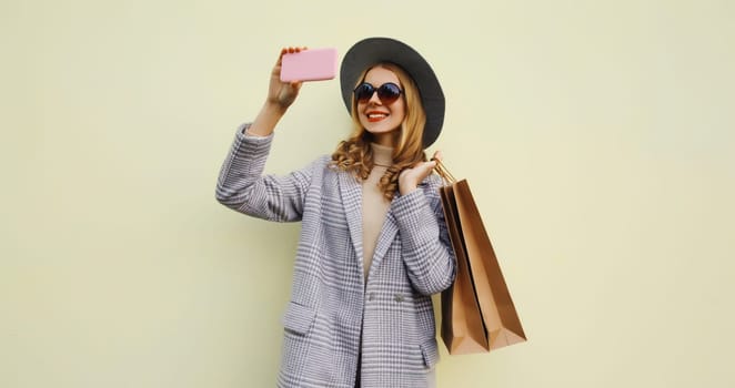 Fashion, shopping, clothing. Beautiful happy young woman taking selfie with phone, stylish smiling girl using smartphone in trendy outfit, hat holding brown shopping bags on white background