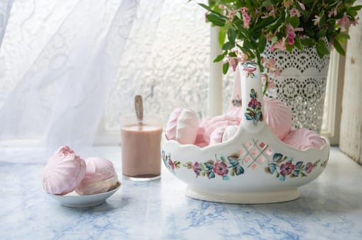 tender still life with full vase of vanilla marshmallows and glass of coffee with milk, next to vase with branches of pink flowers on wooden table, concept country lifestyle,