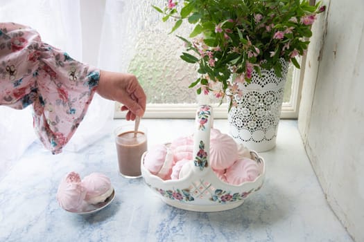 female hand stirring coffee with milk at table with basket of vanilla marshmallows near vase with pink flowers, country life concept,