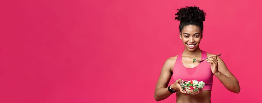 Healthy sporty afro girl eating veggies salad over red studio background, healthy lifestyle concept