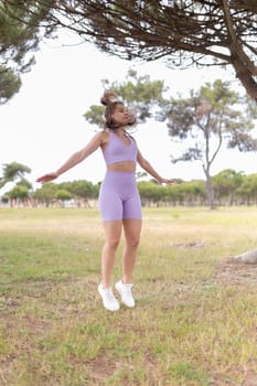 A young woman in a lavender sports bra and shorts performs jumping jacks in a grassy park on a bright day.