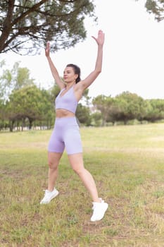 A young woman in athletic wear performs jumping jacks in a grassy park with trees in the background.
