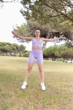 A young woman in a lavender sports bra and shorts performs jumping jacks in a park.