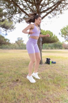 A young woman in purple sportswear jumps while exercising outdoors in a park.