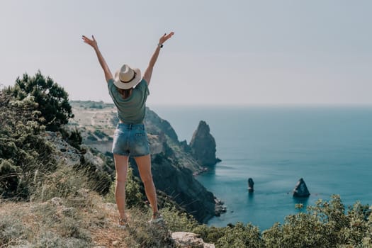 A young woman in a hat stands with her arms raised on a cliff overlooking the ocean.