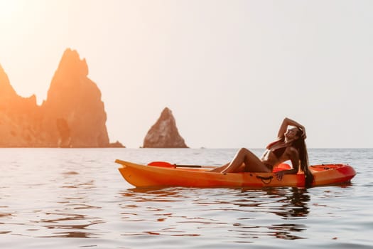 A woman in a bikini reclines on a bright orange kayak in calm water, with dramatic rock formations in the background.