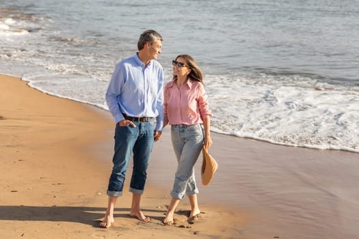 A couple strolls barefoot on the sandy beach, laughing and enjoying each other's company as gentle waves lapping at the shore create a serene atmosphere during sunset.