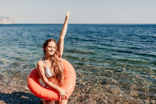 A woman is standing in the ocean with a red inflatable ring. She is smiling and waving