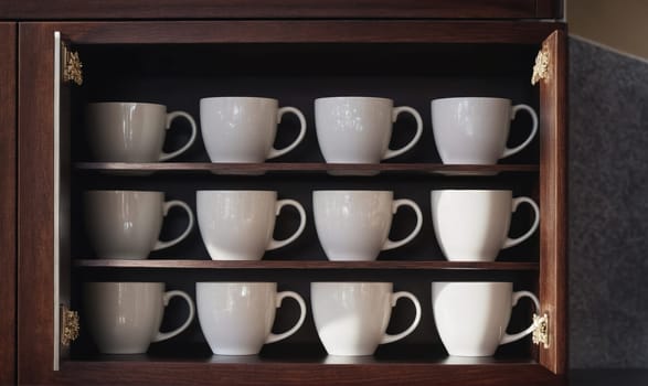 A cabinet door is open, revealing three shelves full of white mugs. The mugs are arranged in rows of four, with their handles facing outward. The cabinet is made of dark wood.