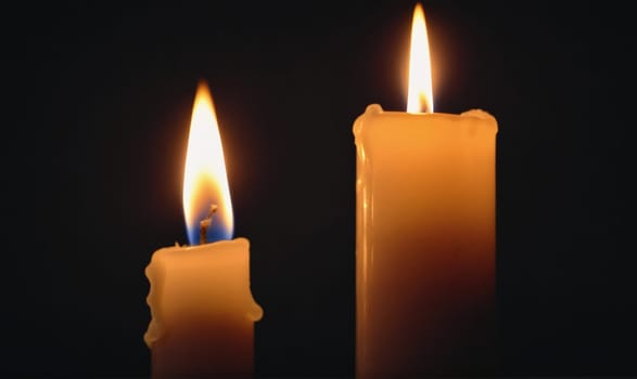 Two candles are lit in a dark room. The candle on the left is smaller and has a blue wick. The candle on the right is taller and has a white wick.