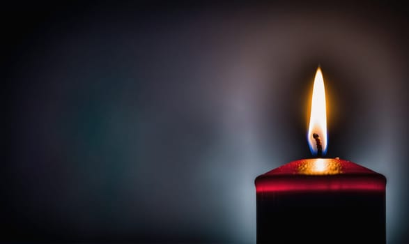 A red candle is lit and burning, casting a soft glow against a dark backdrop. The flame is tall and bright, illuminating the wax.