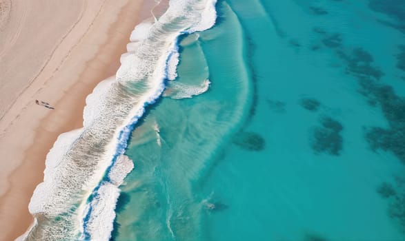 An aerial perspective of a sandy beach, where white waves break on the shore. The water is a vibrant turquoise, with dark patches of coral visible beneath the surface.