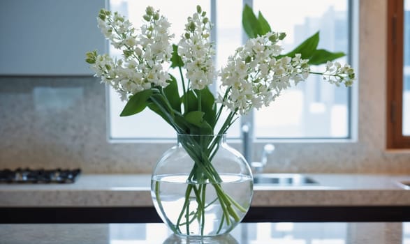 White flowers with green stems are arranged in a clear glass vase, resting on a countertop. The countertop is grey with a black stovetop in the background