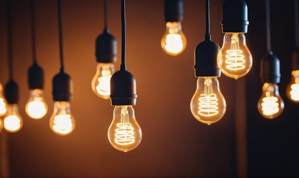 Multiple incandescent light bulbs with a coiled filament are hanging from black cords against a dark brown background. The bulbs are lit and glowing, emitting a warm, yellowish light.