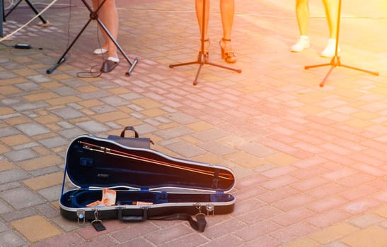 The image shows an open violin case on the ground near the musicians. There is money (tips) in the case. The case is black and made of hard plastic. The musicians are standing in the background.