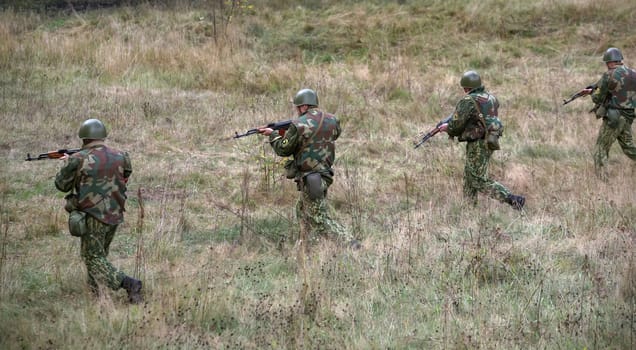 A group of four soldiers dressed in camouflage uniforms maneuvers across a grassy landscape, training for tactical operations in an outdoor setting amidst fall foliage.