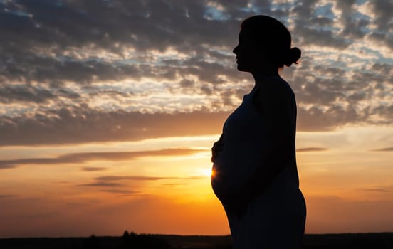 The silhouette of a pregnant woman at sunset with her hands on her belly. The sky is ablaze with color, and the clouds are forming a beautiful pattern. A truly stunning and peaceful scene.