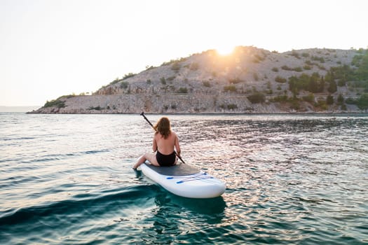 Woman paddle boarding at sunset near a rocky shoreline reflecting golden light on calm waters.