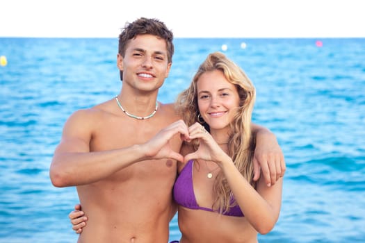 Portrait of happy couple on the beach building a heart with her hands with the sea in the background