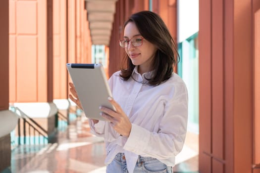 Businesswoman in glasses using digital tablet, standing outdoor dressed white shirt. Female freelancer holding tablet while walk outside
