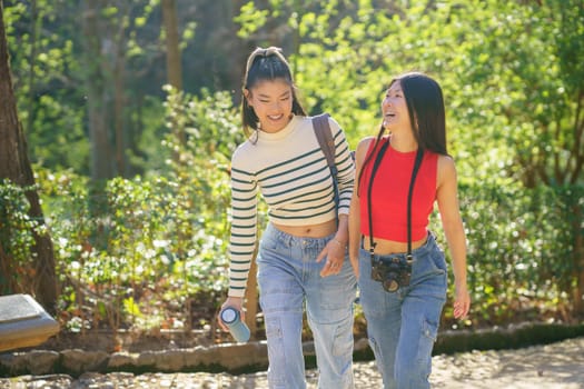 Positive young Asian female friends in casual outfits smiling and speaking about life while strolling together with thermos and camera in green forest of Alhambra