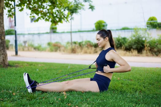 In the park, a young woman prioritizes fitness and wellness by using a resistance band for seated rows. She focuses on strengthening her back muscles as part of an outdoor workout