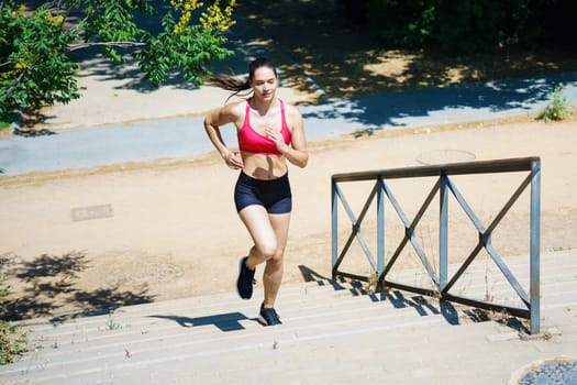 A motivated woman runner in pink sports gear runs up stairs outdoors, displaying determination and athleticism on a sunny day. She prioritizes fitness and health, showing her focus and dedication