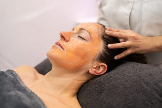 Side view of crop anonymous beautician massaging head of relaxed woman with facial mask during beauty treatment in cosmetician salon