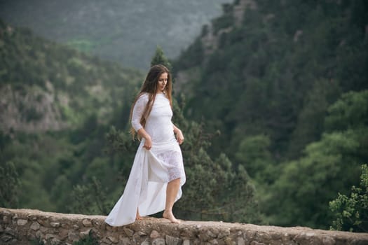 A woman in a white dress is walking on a stone wall. The image has a serene and peaceful mood, as the woman is surrounded by nature and he is enjoying her walk