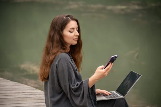 A woman is sitting on the ground with a laptop and a cell phone in her hands. She is using the laptop to browse the internet and the cell phone to communicate with someone